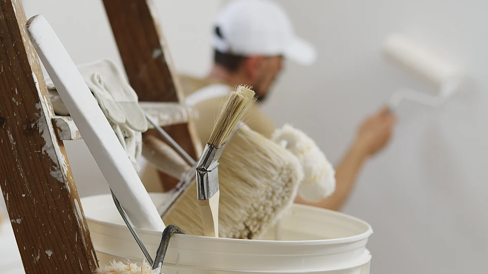 mains du peintre de maison homme décorateur travail de maison à rénover, à l'aide de peinture à rouleaux, d'un seau blanc sur échelle en bois avec des pinceaux en arrière-plan, gros plan