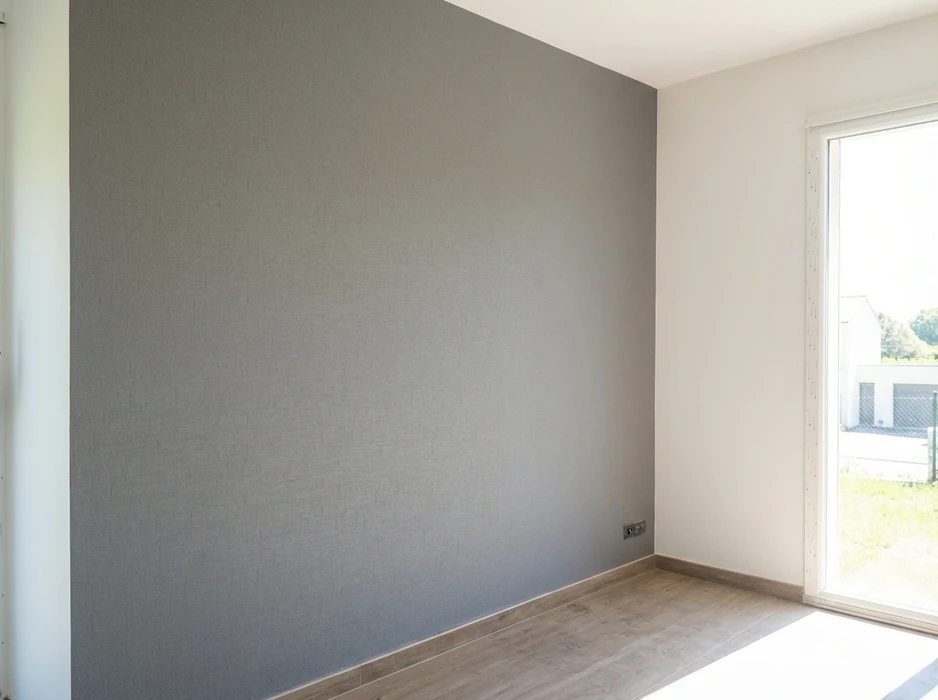 Main décorée d'un gant de protection blanc tenant un pinceau plat appliquant de la peinture grise sur un mur intérieur